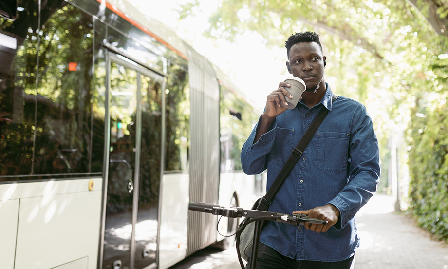 Male entrepreneur with electric push scooter and reusable cup walking by bus